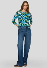 Frau steht mit den Händen in den Taschen, trägt ein langärmeliges Oberteil mit blau-grün-weißem geometrischem Muster und weite blaue Jeans.