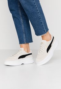 Vaaleat suedesneakersit, joissa on musta sivu raita, valkoinen paksu pohja ja litteät nauhat, yhdistettynä kuluneisiin sinisiin farkkuihin.