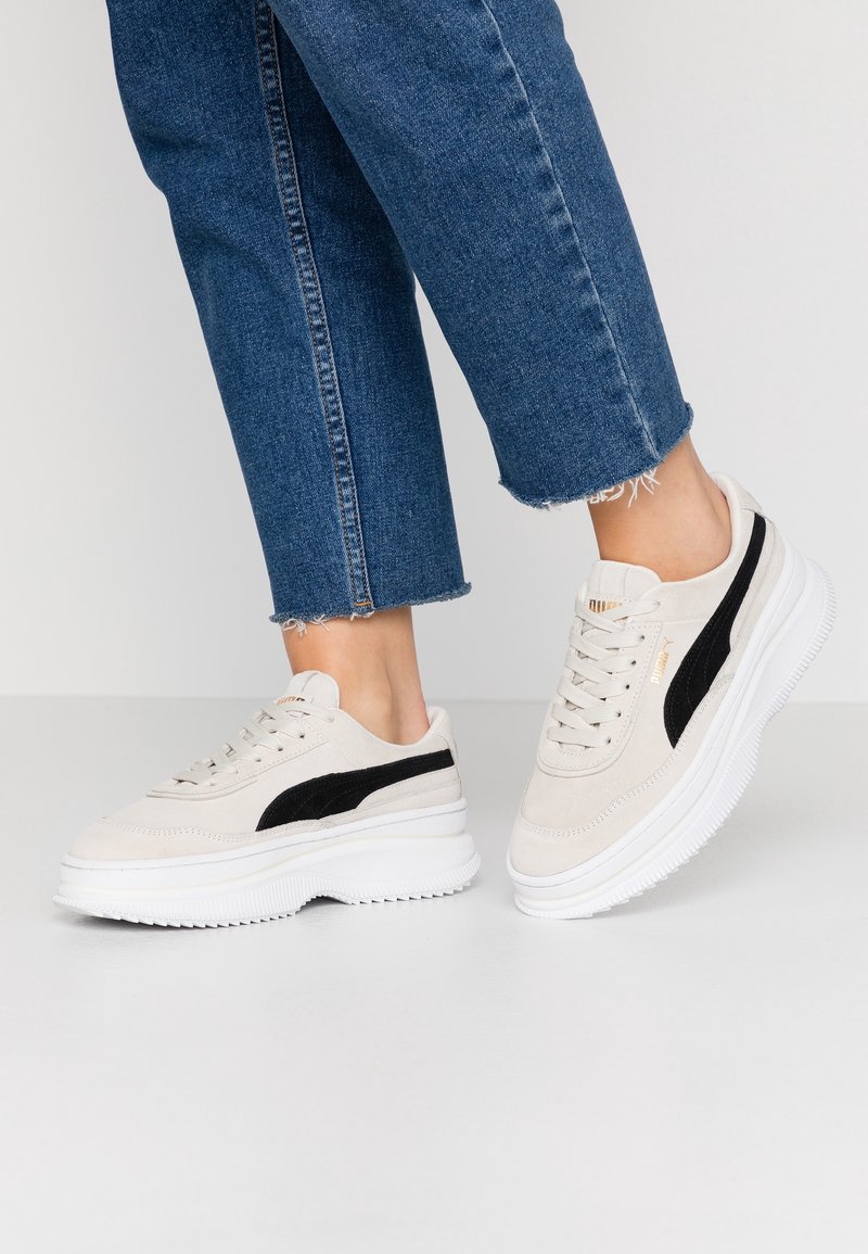 Vaaleat suedesneakersit, joissa on musta sivu raita, valkoinen paksu pohja ja litteät nauhat, yhdistettynä kuluneisiin sinisiin farkkuihin.