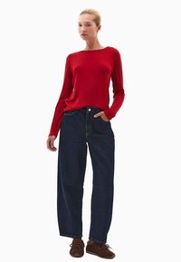 Czerwona bluzka z długim rękawem o luźnym kroju, zestawiona z ciemnoniebieskimi dżinsami typu wide-leg. Brązowe sznurowane buty dopełniają outfit.