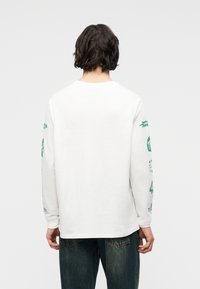 Άτομο με σκούρα μαλλιά μέχρι τον ώμο, που φοράει λευκό πουκάμισο με μακριά μανίκια και πράσινα γραφικά στα μανίκια και σκούρο τζιν, κοιτάζοντας προς τα πίσω.