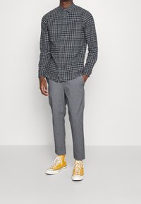 Hombre vestido con una camisa de cuadros gris y negra, pantalones grises y zapatillas de caña alta amarillas, de pie con las manos parcialmente en los bolsillos.