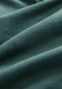 Țesătură verde închis cu o textură moale și netedă, prezentând multiple pliuri și un luciu subtil, indicând un material din bumbac sau o combinație de bumbac.