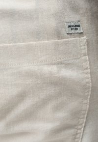 Κοντινό πλάνο σε τσέπη από μπεζ ύφασμα με ορατές ραφές και μια μικρή λευκή ετικέτα στην πάνω δεξιά γωνία που αναγράφει "JACK & JONES EST. 1990".