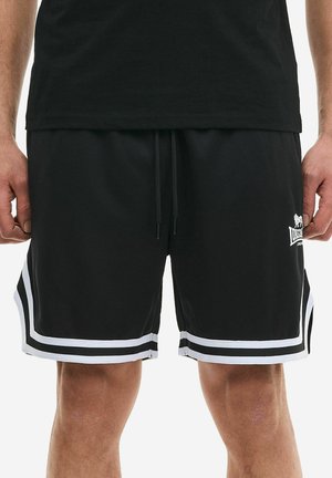Homme portant un short de sport noir avec des bandes blanches à l'ourlet et un petit logo blanc sur la jambe gauche, assorti à un t-shirt noir.
