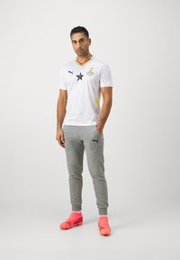 Hvid fodboldtrøje med sorte detaljer og farverne fra Ghanas flag. Grå sweatpants, lyserøde fodboldstøvler og et sort stjerne-logo vist fremtrædende.