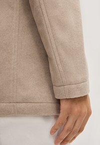 Beige wollen blendjacket met een gladde textuur, op maat gemaakte ontwerp en zichtbare stiksels langs de manchetten en de zoom. Hand ligt op de jas.