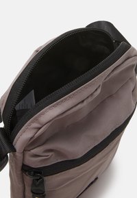 Brown fabric crossbody bag with a black zipper and strap. Lined interior shows pockets and label, featuring smooth texture and compact design.
