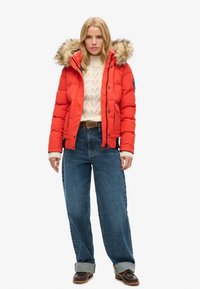 Rode pufferjack met een nepvelour-lined capuchon; gequilt textuur en ritssluiting aan de voorkant. Gecombineerd met een crèmekleurige gebreide trui en losse blauwe jeans.