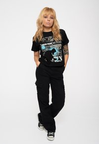 Black cotton t-shirt with graphic print featuring "Linkin Park Meteora." Paired with black cargo pants and black sneakers. Model poses with hands in pockets.