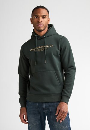 Man met een donkergroene hoodie met gele "Petrol Industries & Co." tekst en een blauwe spijkerbroek, staand tegen een effen lichte achtergrond.