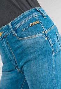 Jean bleu clair en denim avec des accents en métal doré et une étiquette de marque visible. Comprend des poches standard et des détails de couture.