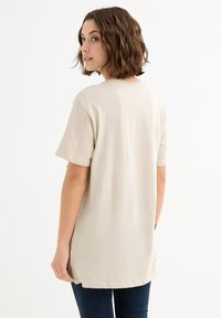Camiseta de algodón beige con mangas cortas, corte relajado, escote redondo y bajo ligeramente asimétrico, combinada con jeans azul oscuro.