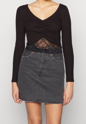 Vrouw draagt een zwarte, geplooide top met lange mouwen en kantdetail, gecombineerd met een hooggetailleerde, vervaagde zwarte denim minirok.