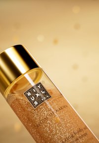 Bouteille en verre transparent avec un couvercle doré, remplie d'huile corporelle dorée scintillante. Logo et texte en évidence sur le devant. Fond doré.