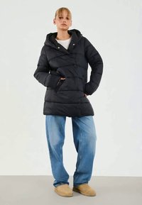 Giacca a vento nera con cappuccio, caratterizzata da cuciture diagonali, due tasche laterali e una vestibilità comoda, abbinata a jeans blu larghi e scarpe beige.