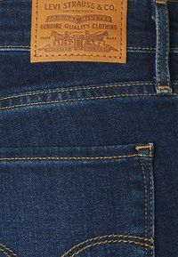 Vaqueros de denim azul oscuro con una etiqueta de cuero marrón con texto e imágenes en relieve, costuras doradas y dos bolsillos traseros.