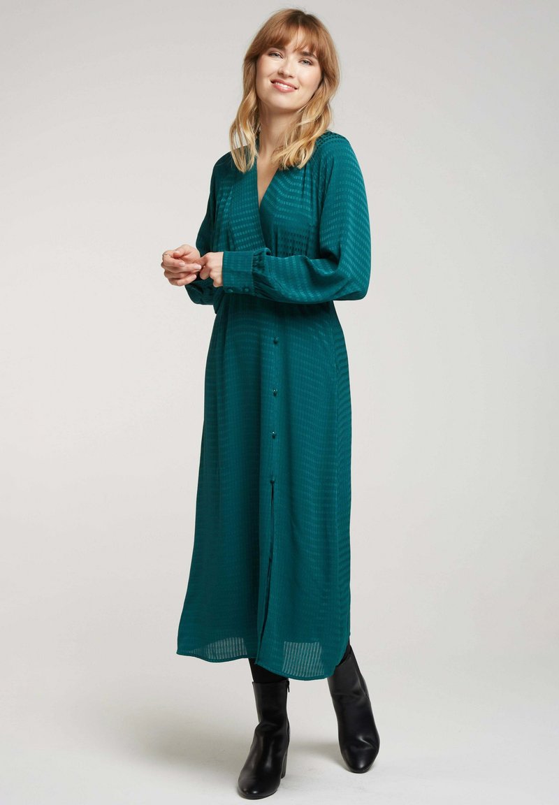 Robe longue teal à manches longues avec un décolleté croisé, un bouton à l'avant et des rayures verticales texturées. Portée avec des bottes noires montant jusqu'aux genoux.