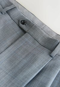 Pantalon sur mesure en tartan gris avec bouton à la taille, passants de ceinture et plis sur fond blanc.
