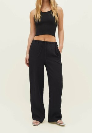 Pantalones anchos negros hechos de una tela texturizada con una cinturilla elástica y cordón. Combinados con un top corto negro ajustado y sandalias plateadas.