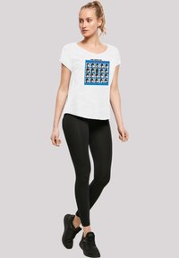 Weißes T-Shirt mit kurzen Ärmeln, das einen blauen Grafikdruck mit Gesichtsdarstellungen zeigt, kombiniert mit schwarzen Leggings und schwarzen Sport-Schuhen.