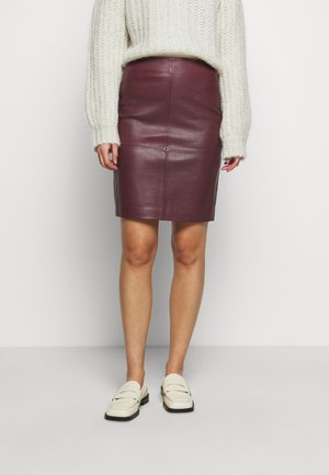 Kvinde iført en burgundy læder nederdel til knæet, cream chunky striktrøje og hvide loafers, stående mod en ensfarvet lys baggrund.