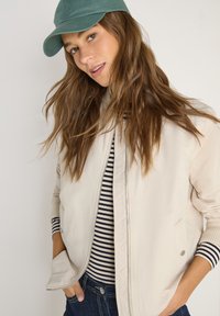 Jeune femme aux longs cheveux bruns portant une casquette verte, une veste beige et un haut rayé noir et blanc, les mains dans les poches.