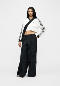 Νεαρή γυναίκα που φοράει λευκή κοντή φούτερ Adidas με μαύρες ρίγες και φαρδιά μαύρη φόρμα Adidas, στέκεται με το ένα χέρι στη μέση.