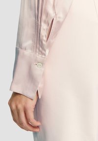 Blusa di satin rosa chiaro con polsino con bottone e dettagli plissettati. La superficie liscia e la forma sartoriale enfatizzano l'eleganza.