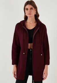 Bordo per didelis hoodi paltas su užtrauktuku priekyje ir raišteliais. Savybės: tekstūruota medžiaga ir ilgomis rankovėmis. Dėvimas ant juodo trumpaaukščio.