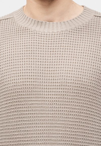 Gros plan d'une personne portant un pull beige en maille gaufrée avec un col rond côtelé.