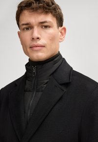 Zwarte jas met een gestructureerd ontwerp, voorzien van een ritskraag en een gladde textuur. Gelaagd over een zwart donsjacket.