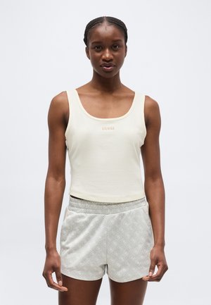 Femme aux cheveux tressés portant un débardeur crème côtelé avec le logo "GUESS" et un short gris clair à motifs, debout devant un fond uni.