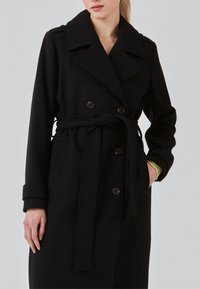 Woman wearing a black belted double-breasted coat with buttons and a popped collar, one hand in pocket, against a plain light background.