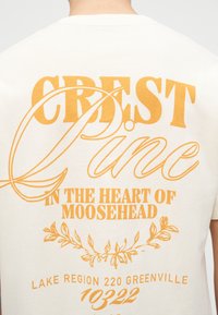 Πίσω μέρος λευκού T-shirt με μεγάλο πορτοκαλί κείμενο που γράφει "CREST Dune, στην καρδιά του Moosehead," διακοσμητικά φύλλα και λεπτομέρειες τοποθεσίας.