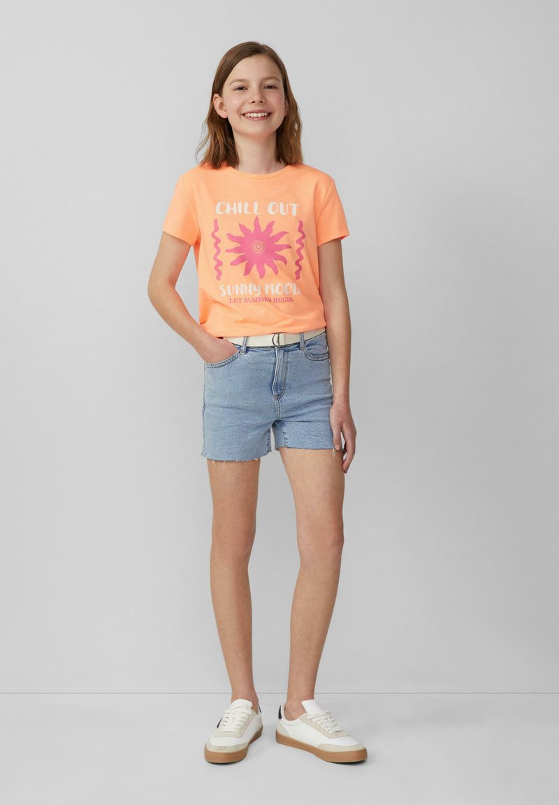 Helles orange Baumwoll-T-Shirt mit pinkem Sonnen-Graphic und Text, kombiniert mit hellen Jeansshorts. Weiße Sneakers mit braunen Akzenten.