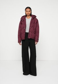Burgundijas krāsas quilted puffer jakas ar kapuci, kombinēta ar gaiši pelēku blūzi un melniem platām kājām biksēm. Iezīmes snap aizdare un teksturēts dizains.