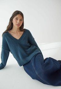Teal stickad tröja med bred V-ringning, sänkta axlar och ribbad nederkant, ihop med en mörk denimkjol. Slät textur.