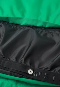 Grüner Stoff mit einem schwarzen Innenfutter; verfügt über einen Reißverschluss und einen texturierten Riemen mit geprägten Größenangaben.