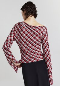 Persona con capelli ricci corti che indossa una maglietta a maniche lunghe a quadri rossi, bianchi e neri con scollatura asimmetrica, pantaloni neri, di spalle.