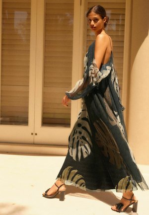 Robe fluide et longue en tissu transparent, avec une base sombre ornée de grands motifs de feuilles vertes et beiges. Accentuée par des bretelles spaghetti.
