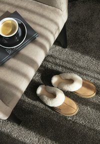 Tawny, met bont gevoerde slippers op een grijs tapijt naast een beige bank met zwarte poten, bovenop een boek en een zwart koffiekopje met espresso.