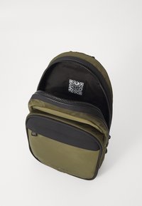 Olive green backpack with black accents, zip closure, and front pocket. Interior features a QR code label. Material is durable and textured.
