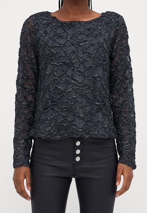 Schwarzes transparentes Langarmoberteil mit wellenförmigen Strukturen, kombiniert mit hoch taillierten schwarzen Hosen mit drei silbernen Knöpfen.