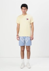 Lys gul bomull T-skjorte med et lite flerfarget logo, kombinert med blå mønstrede shorts og hvite joggesko.