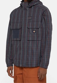 Man som bär en marinblå, röd och vit rutig huva-jacka med flera fickor fram och brända orange cargobyxor.