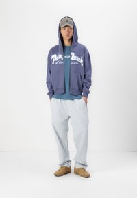 Mørkeblå zip-up hættejakke med "Palm Beach" grafik, lyseblå t-shirt under, lysegrå løse bukser og brune sko.