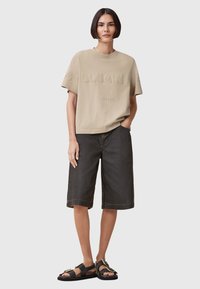 T-shirt in cotone beige con logo in rilievo, abbinato a pantaloni shorts in denim neri larghi e sandali neri con dettagli dorati.