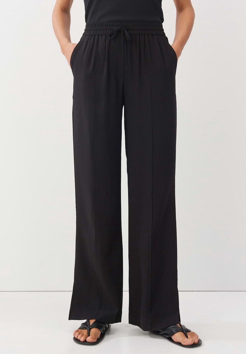Personne portant un pantalon noir ample avec une taille élastique et des sandales noires à lanières croisées sur les orteils, debout les mains dans les poches devant un fond uni.