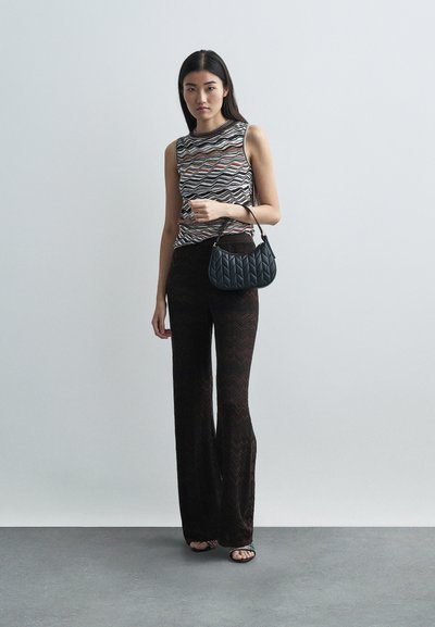 Γυναίκα στέκεται δίπλα σε λεία τοιχογραφία, φορώντας αμάνικο εμπριμέ τοπ, μαύρο παντελόνι με ζιγκ-ζαγκ σχέδιο, κρατώντας μικρή τσαντάκι από καπιτονέ σε μαύρο χρώμα και φορώντας ανοιχτά παπούτσια με τακούνι.